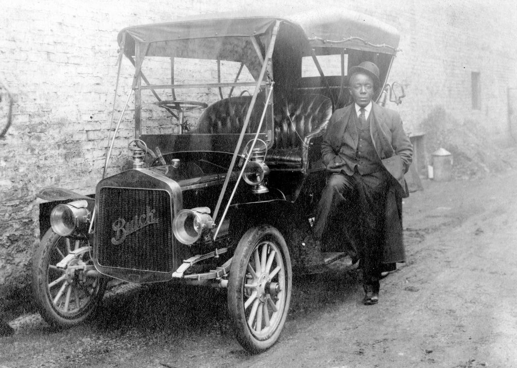 Black Wall Street - Full length shot of African American man leaning against a Buick car parked against a building, wearing a suit and hat tilted on his head, neutral facial expression, 1920.