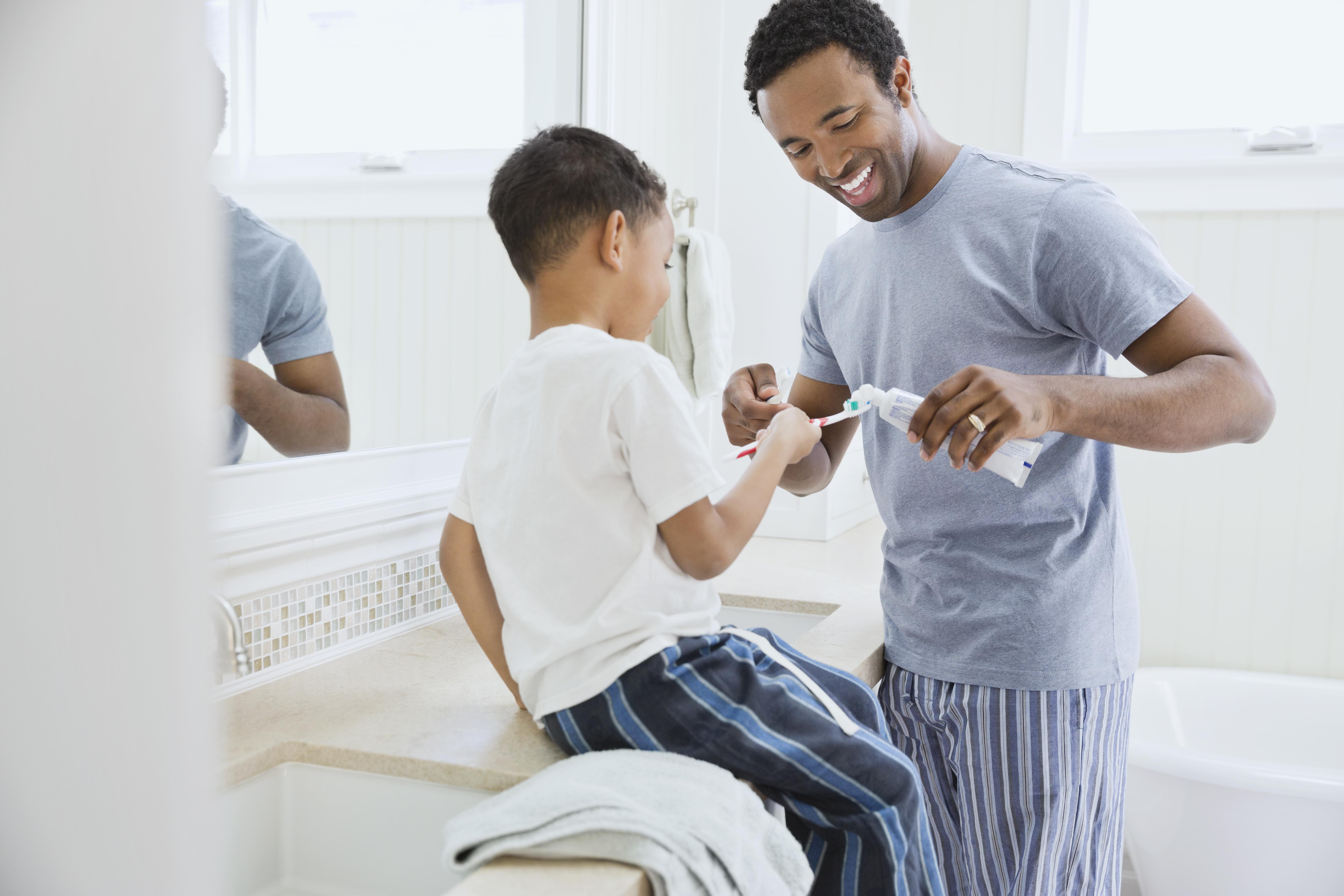 Father squeezing toothpaste on son's toothbrush in domestic bathroom